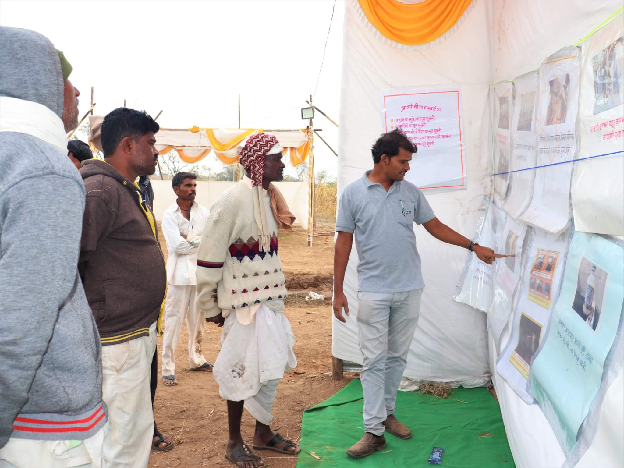 A group of people who are gathered in one of the informational poster galleries watches as an Animal Rahat team member elaborates on the content of one of the posters.